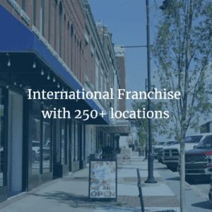 Street view of a storefront with a sign reading "We Are Open" on a sidewalk. Overlaid text says "International Franchise with 250+ locations.